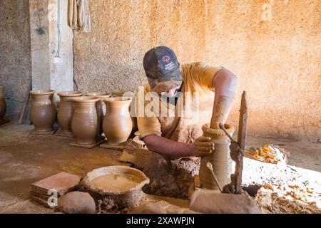 Un uomo lavora su un pezzo di ceramica in una stanza con molti altri pezzi. L'atmosfera è calma e concentrata, mentre l'uomo modella con cura l'argilla Foto Stock