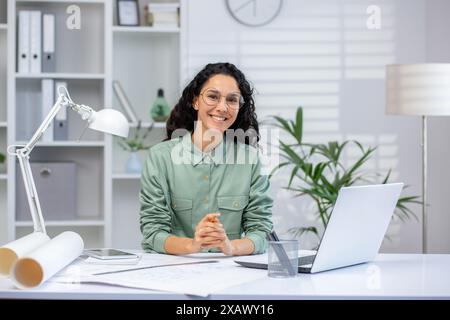 Donna sicura di sé seduta alla scrivania con un notebook, che guarda la fotocamera con un sorriso, in un ambiente moderno e luminoso. Foto Stock