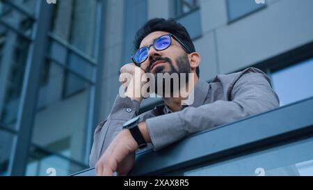 Triste annoiato uomo etnico arabo indiano stanco datore di lavoro uomo d'affari attendono la riunione sul balcone della città ufficio edificio controllare il tempo di attesa su orologio da polso Foto Stock