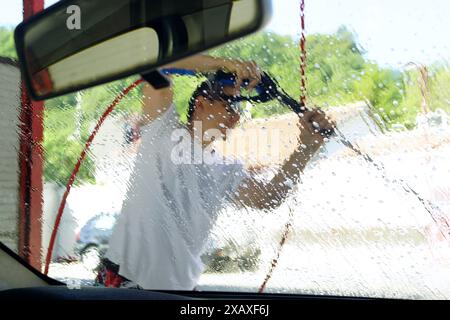 Vista dal sedile del passeggero di un uomo che pulisce il cofano e il parabrezza di un'auto utilizzando una pistola ad alta pressione. Foto Stock