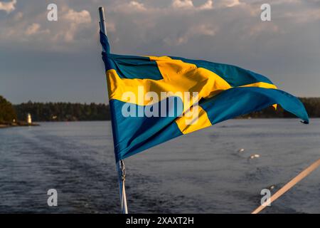 La bandiera svedese sventola dalla poppa della nave da crociera alla luce della sera, con un piccolo faro visibile su un’isola di un arcipelago sullo sfondo. Karlsuddsvägen, Vaxholms kommun, Stoccolma, Svezia. Foto Stock