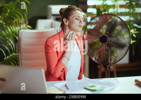 Ambiente di lavoro sostenibile. donna moderna di 40 anni lavoratrice al lavoro in giacca rossa con ventilatore elettrico e tovagliolo che soffrono di caldo estivo. Foto Stock