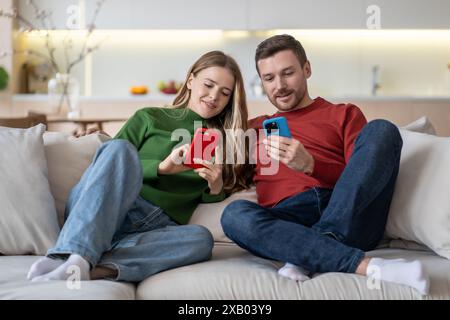 Uomo e donna felici seduti in salotto con sorrisi che guardano allegramente gli schermi dei telefoni cellulari. Foto Stock
