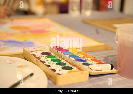 Pitture e materiali da disegno su un tavolo in un laboratorio d'arte per bambini. Bambini impegnati in attività creative guidate dal loro insegnante. Foto Stock