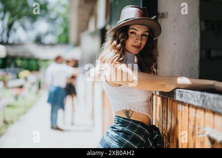 Giovane donna in un elegante cappello appoggiata su una recinzione presso una scuderia di cavalli in una giornata di sole Foto Stock