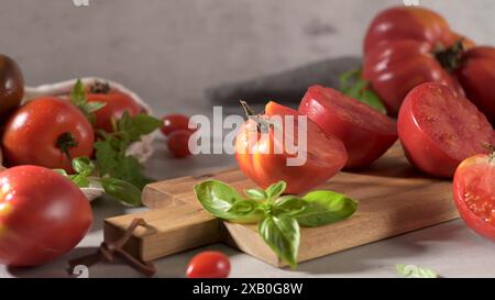 Una varietà di pomodori rossi, interi e tagliati a metà, su un asse di legno, guarniti con basilico. Foto Stock