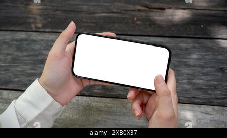 Immagine ravvicinata della mano di una donna che tiene in mano uno smartphone con un modello a schermo bianco in posizione orizzontale su un tavolo di legno in un giardino. la gente è una Foto Stock