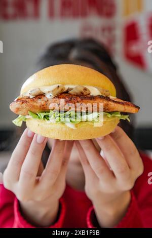 Donna che tiene in mano un succoso e tenero hamburger di brioche di pollo grigliato con le dita davanti al viso che si nasconde dietro. Foto Stock