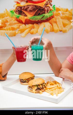 Le mani di due donne fanno il tifo per sorseggiare bibite alla limonata con hamburger e patatine fritte sul tavolo. Foto Stock