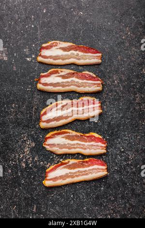 Immagine ravvicinata di cinque strisce di bacon crude disposte su una superficie nera e ruvida. Vista dall'alto. Foto Stock