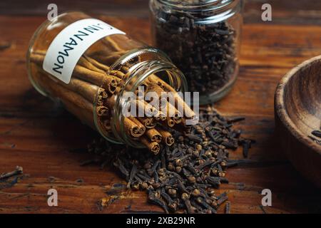 Cannella e chiodi di garofano fuoriescono dal vaso di vetro delle spezie su un tavolo di legno, concetto di utilizzo di spezie in cucina Foto Stock