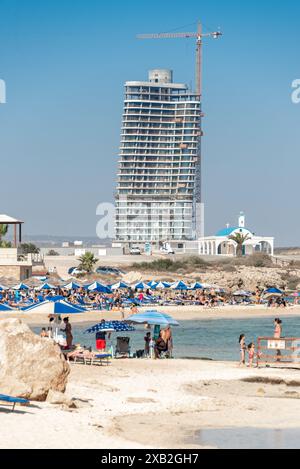 Ayia Napa, Cipro - 2 luglio 2022: Turisti che si rilassano sulla spiaggia di Ayia Thekla con un alto edificio incompiuto di Ayia Napa Marina e gru da costruzione Foto Stock