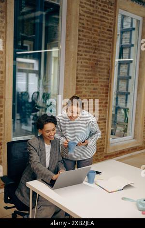 Due colleghi donne concentrate che lavorano insieme al progetto e utilizzano un laptop seduto in ufficio Foto Stock