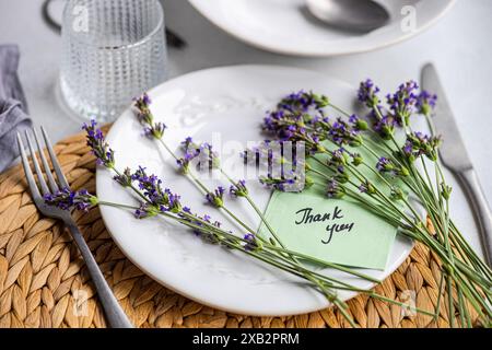 Un tavolo estivo splendidamente allestito con un piatto bianco adornato da vivaci fiori di lavanda e un biglietto di ringraziamento, che simboleggia l'eleganza Foto Stock