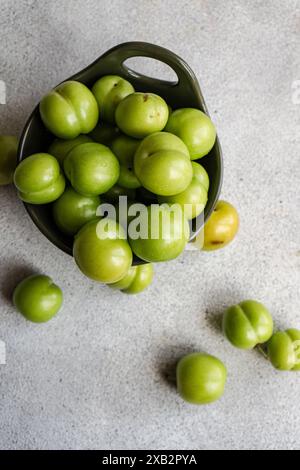 Una ciotola piena di prugne verdi organiche mature si trova su una superficie grigia testurizzata, con alcune prugne sparse intorno ad essa, questi frutti sono popolari in vari medi Foto Stock