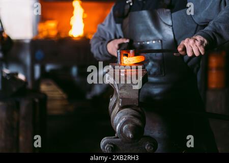 Un fabbro corto e irriconoscibile forma un pezzo di metallo riscaldato su un'incudine con un martello in una fucina tradizionale. Foto Stock