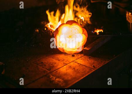 Un pezzo luminoso di metallo a forma di fiore irradia calore nella fucina di un fabbro, con fiamme che leccano l'aria intorno ad essa. Foto Stock