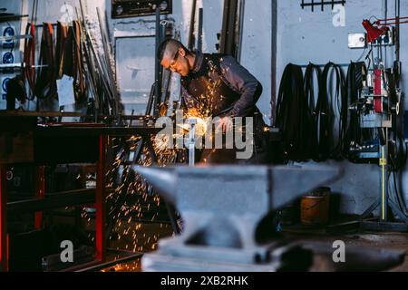 Un fabbro maschio è incentrato sulla molatura di un pezzo di metallo, con scintille luminose che volano in officina. Foto Stock