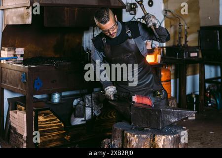 Un fabbro maschio sta modellando un pezzo di metallo caldo rosso su un'incudine con un martello in un ambiente tradizionale da officina. Foto Stock