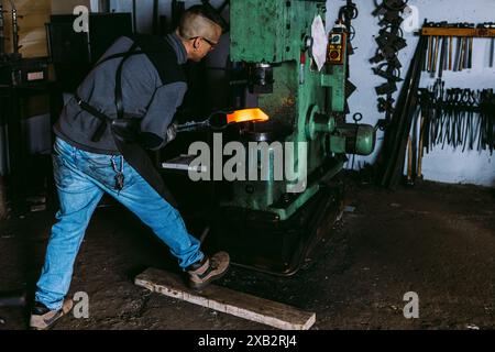 Fabbro maschio anonimo che forgia metallo caldo con un martello elettrico in un'officina scura. Foto Stock