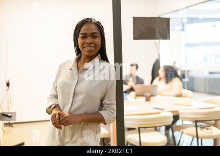 Una donna sorridente professionista si trova in primo piano in uno spazio ufficio mentre il suo team lavora a un tavolo dietro di lei, illustrando un cowo collaborativo Foto Stock