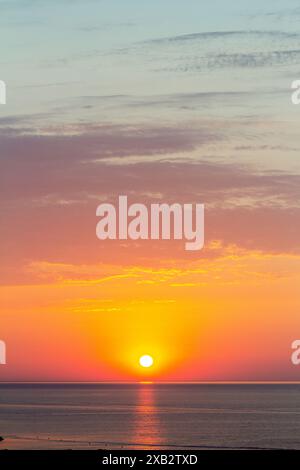 Una vista serena del sole che tramonta sull'oceano, proiettando un brillante bagliore arancione e rosa sul cielo, le acque calme riflettono la luce, creando un p Foto Stock