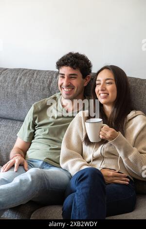 Una giovane coppia allegra siede comodamente su un divano grigio, condividendo un momento di luce con una tazza di caffè in un accogliente soggiorno Foto Stock