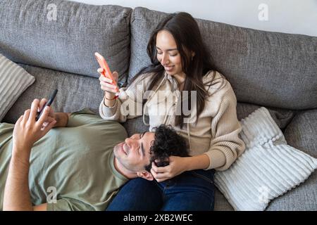 Un uomo e una donna comodamente sdraiati sul divano, sorridenti mentre utilizzano il proprio smartphone in un ambiente accogliente, dimostrando un momento di disinvoltura Foto Stock