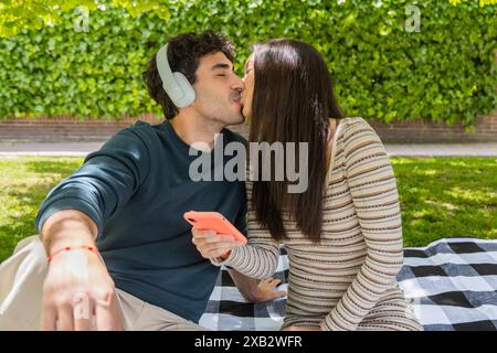 Una giovane coppia si siede su una coperta a scacchi in un parco lussureggiante, condividendo un bacio l'uomo indossa le cuffie e tiene in mano uno smartphone, mentre la donna sorride e. Foto Stock