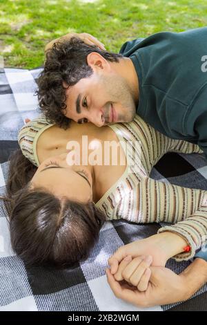 Una giovane coppia gode di un momento tranquillo insieme, sdraiati su una coperta da picnic in un parco, si tengono per mano e condividono uno sguardo amorevole, circondati dal verde Foto Stock