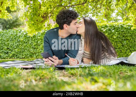 Un giovane uomo e una donna condividono un bacio tenero mentre si siedono su una coperta a scacchi in un vivace parco verde, la coppia gode di un momento sereno, con uno smar Foto Stock