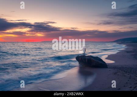 Silhouette solitaria di una persona irriconoscibile in piedi su una roccia che guarda il vibrante tramonto sulla spiaggia di Atlanterra a Cadice. Foto Stock
