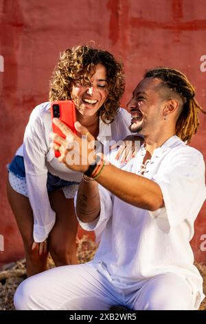 Una coppia allegra si gode una calda giornata di sole scattando selfie con uno smartphone rosso i loro sorrisi riflettono i momenti felici condivisi, vestiti casualmente di bianco Foto Stock