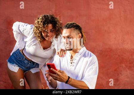 Un uomo e una donna gioiosi stanno vicini, condividendo contenuti su uno smartphone, su una vivace parete rossa, mettendo in risalto i loro outfit casual Foto Stock