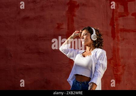 Una giovane donna con una camicetta bianca corta e jeans blu ama ascoltare la musica attraverso le cuffie bianche, appoggiandosi su una vivace parete rossa con la luce del sole castin Foto Stock