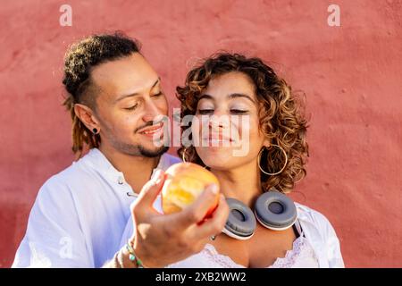 Un giovane uomo e una donna sorridono amorevolmente mentre condividono una grande mela, in piedi contro una vivace parete rosa la donna indossa un top bianco e le cuffie Foto Stock