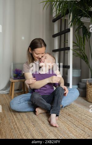 Una madre amorevole e suo figlio gioioso condividono un abbraccio e una risata mentre si siedono su un accogliente pavimento tappezzato a casa, vicino a un acquario, incarnando una calda, familia Foto Stock