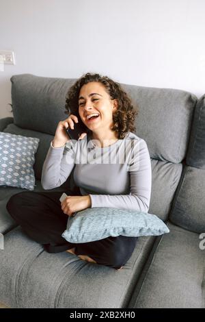 Una donna ride gioiosamente durante una conversazione telefonica, seduta su un comodo divano con un cuscino in grembo. Foto Stock