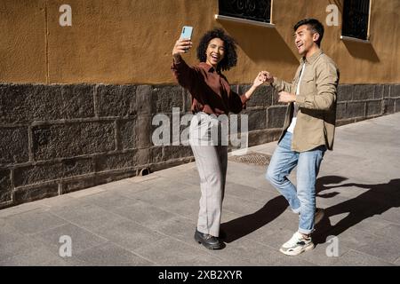 Un momento di allegria mentre una coppia multietnica si fa un selfie e si tiene per mano in una giornata di sole a Madrid, Spagna. L'uomo cinese e la donna ispanica condividono una j Foto Stock