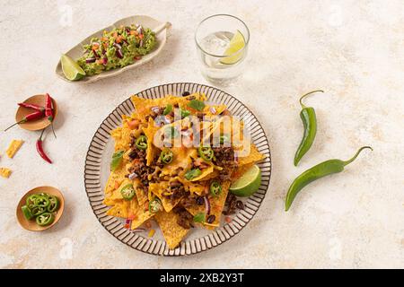 Vista dall'alto di un piatto di nachos abbondantemente condito con formaggio fuso, manzo macinato, jalapenos ed erbe fresche, abbinato a guacamole e lime su un leggero bicchiere Foto Stock