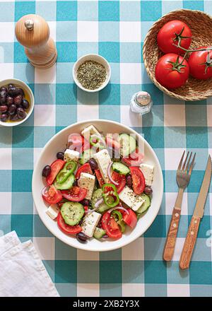 Una vivace ciotola di insalata greca con cubetti di formaggio feta, pomodori a fette, cetrioli e olive, posta su una tovaglia a scacchi blu e bianca E su Un cestino Foto Stock