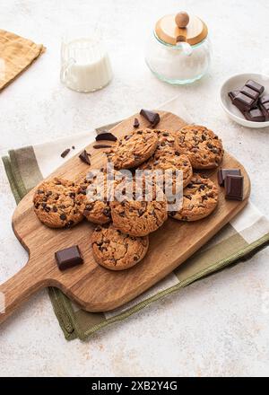 Biscotti con gocce di cioccolato appena sfornati disposti su un piatto da portata in legno, accompagnati da una caraffa di latte e pezzi di cioccolato fondente Foto Stock