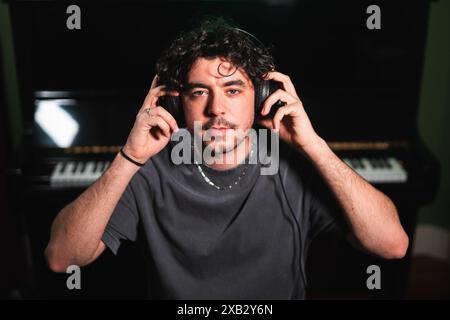 Un giovane uomo concentrato regola le cuffie in uno studio di registrazione poco illuminato, pronto a mixare i brani sul sintetizzatore dietro di lui Foto Stock