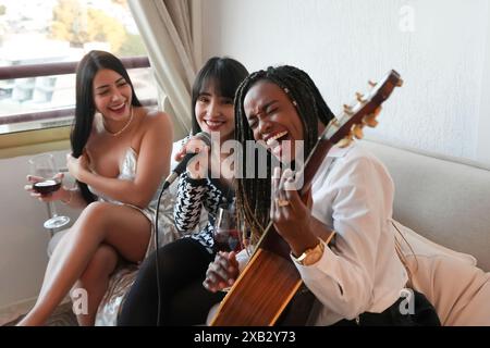 Tre amici condividono un momento di gioia in una stanza accogliente, uno suona la chitarra e canta, mentre gli altri ascoltano e si godono il vino, ridendo insieme in una celebrità Foto Stock