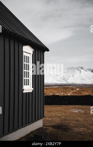 Un caratteristico cottage islandese tradizionale nero con finestre con finiture bianche, si erge in netto contrasto con lo sfondo delle montagne innevate sul Foto Stock