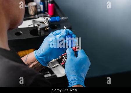 Primo piano di un irriconoscibile tatuatore che indossa i guanti blu, creando una macchina per tatuaggi con strumenti sfocati sullo sfondo. Foto Stock
