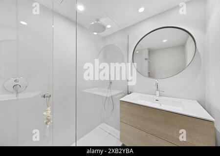 Bagno moderno con combinazione di colori bianchi, specchio arrotondato, lavabo a parete e box doccia in vetro Foto Stock