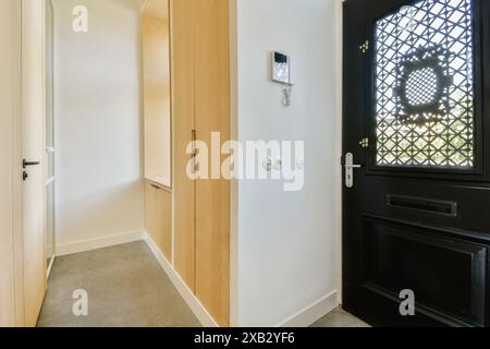 Un luminoso e invitante ingresso moderno con una porta di sicurezza nera con una finestra decorativa a motivi geometrici, fiancheggiata da pannelli di legno e una wa bianca Foto Stock