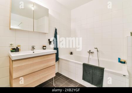 Un bagno elegante e moderno con piastrelle bianche immacolate, un lavandino in legno con lavandino, un grande specchio e una vasca bianca impreziosita da asciugamani color tè scuro Foto Stock