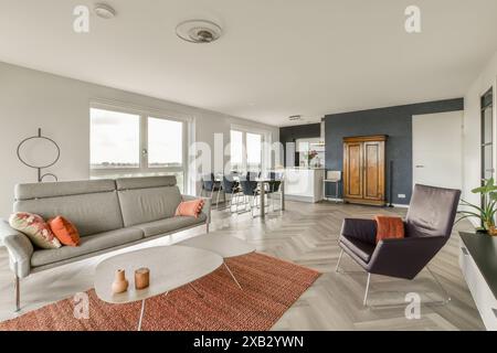 Un soggiorno arioso e moderno con divano grigio, sedia contemporanea, tavolino rotondo e cucina integrata con sgabelli da bar. Foto Stock